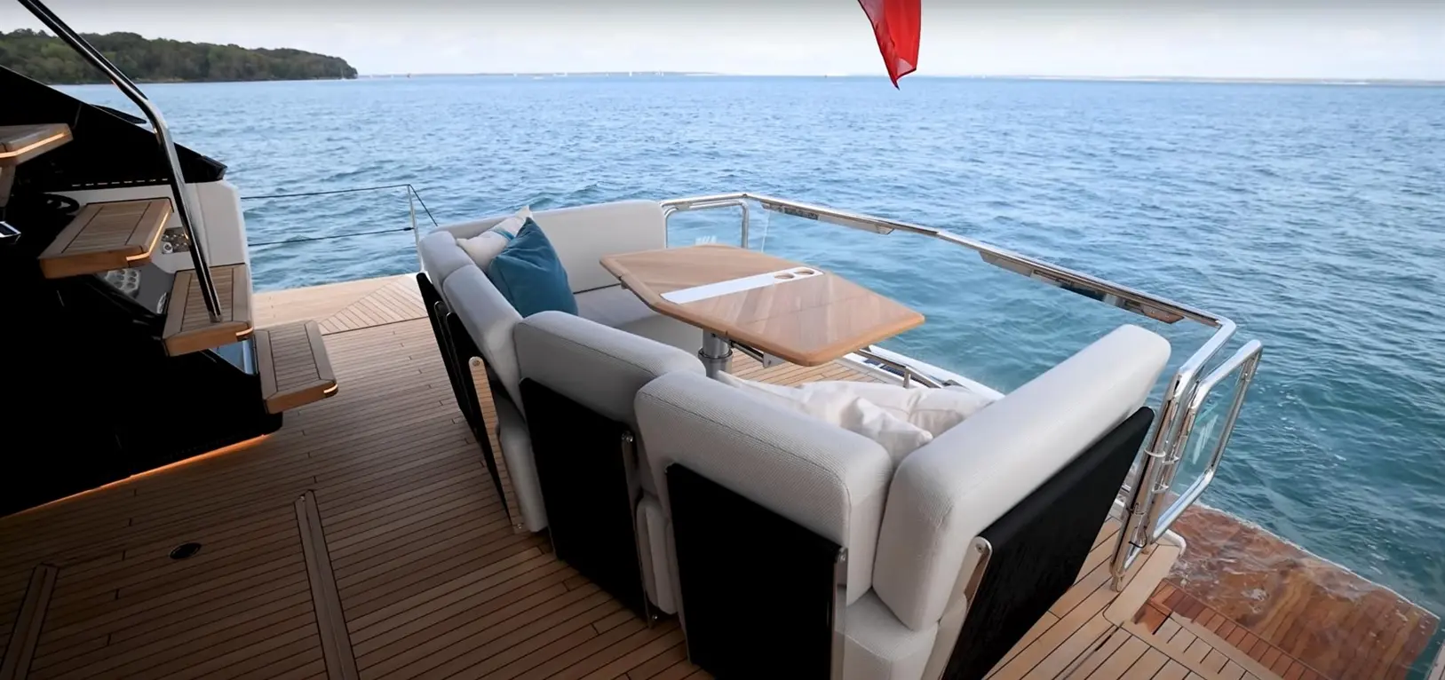 A luxury yacht's deck features a plush seating area with a wooden table, overlooking calm blue waters and greenery in the background.